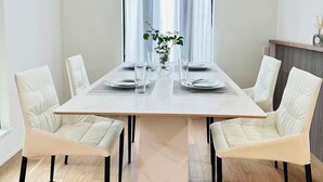 Ruokailuhuoneessa on suorakulmainen pöytä, kuusi valkoista, pehmustettua tuolia, riippuvalaisin ja ikkuna, jossa on läpikuultavat verhot.