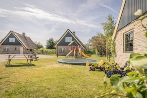 Ein Spielplatz mit Rutsche und Trampolin, ein Picknicktisch und zwei Häuser mit Backsteinfassade.