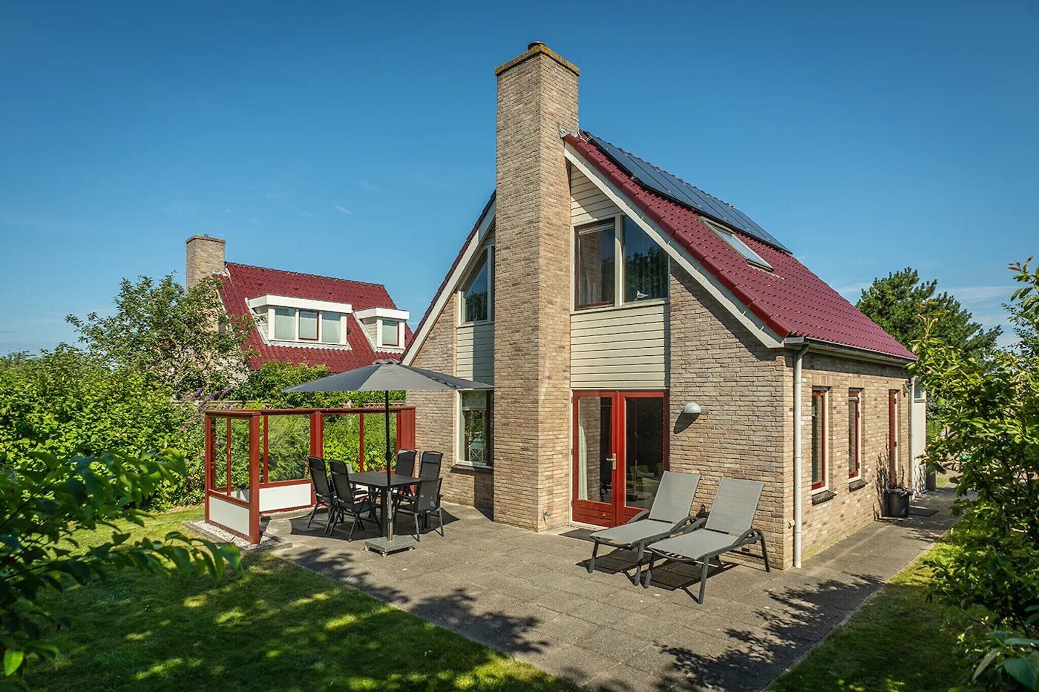 A house with a sloped roof, a chimney, a glass enclosure with chairs, and a patio area.
