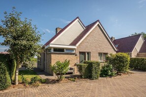 Uma casa de dois andares com fachada de tijolos, telhado inclinado e um jardim frontal com sebes e uma árvore.