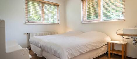 Quarto com cama, mesa de cabeceira, radiador e duas janelas com persianas.