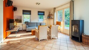 Ein geräumiges Wohnzimmer mit einer Couch, Sesseln, einem Couchtisch und einem Fernseher. Zwei Fenster mit Jalousien, ein Kamin und ein Bücherregal sind ebenfalls vorhanden.