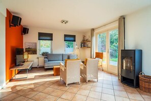 Ein geräumiges Wohnzimmer mit einer Couch, Sesseln, einem Couchtisch und einem Fernseher. Zwei Fenster mit Jalousien, ein Kamin und ein Bücherregal sind ebenfalls vorhanden.