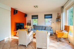 Un salon comprenant un canapé, deux fauteuils, un téléviseur et une fenêtre avec des stores.