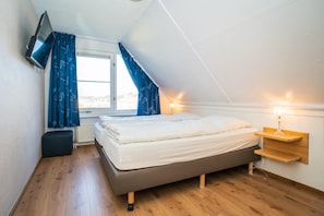 A bedroom with a bed, a TV mounted on the wall, a window with curtains, and a bedside table with a lamp.