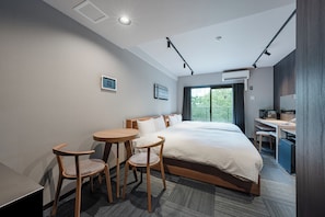 Habitación de hotel con una cama grande, dos sillas, una mesita, un escritorio con una computadora y un ventanal con vistas al verdor.