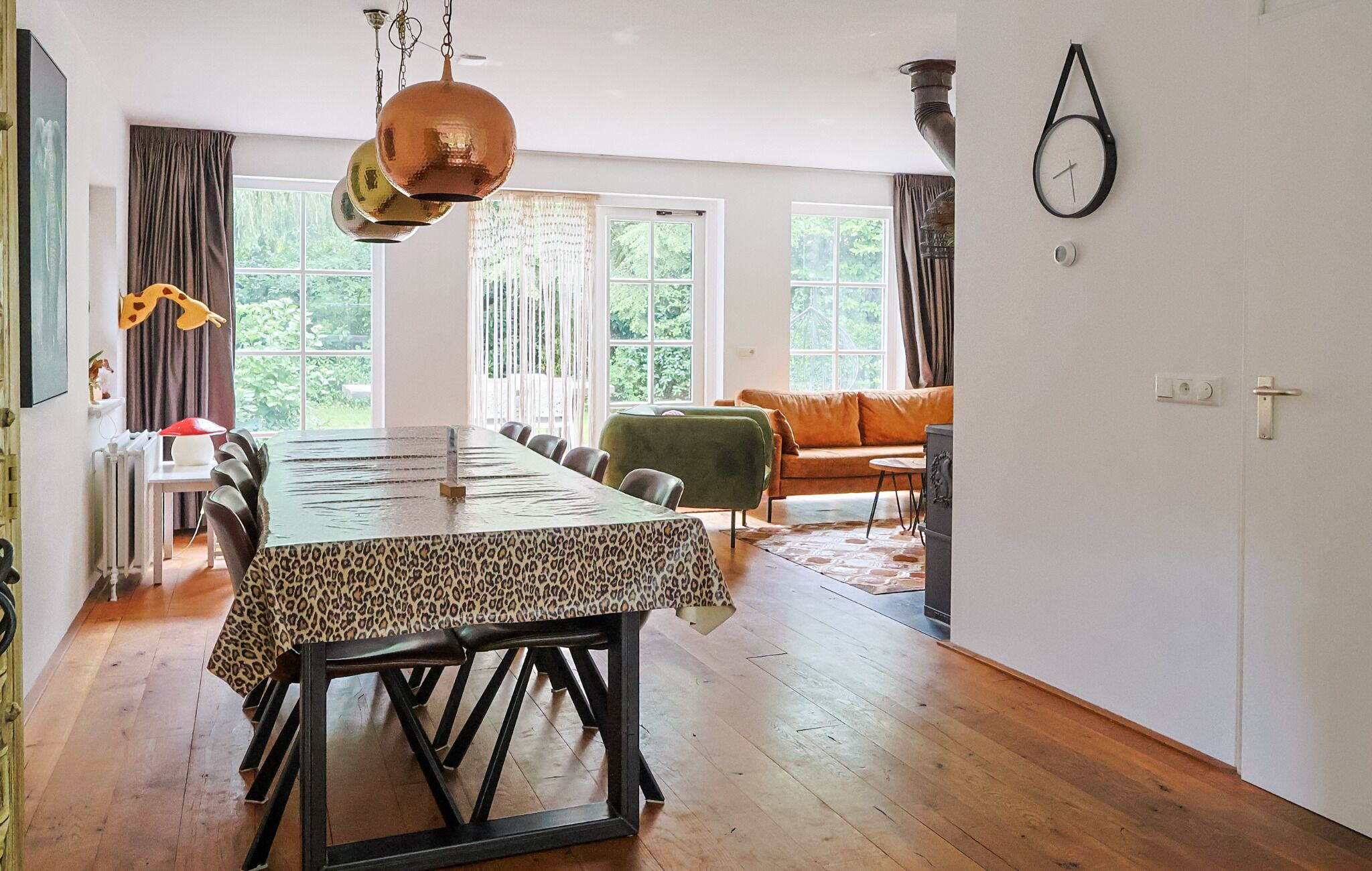 A dining area with a table, chairs, and a chandelier. There are windows with curtains, a sofa, and a clock on the wall.