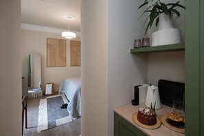 A hotel room with a bed, a bedside table with a lamp, a mirror, a wall-mounted shelf with decorative items, and a small plant. - Family Room, The Kodrian (Athens)