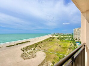 A beachfront view with a sandy beach, a grassy area with a curved path, and a high-rise building in the background. - Harbour Light Towers 1702 Beachfront and Renovated (Clearwater Beach)
