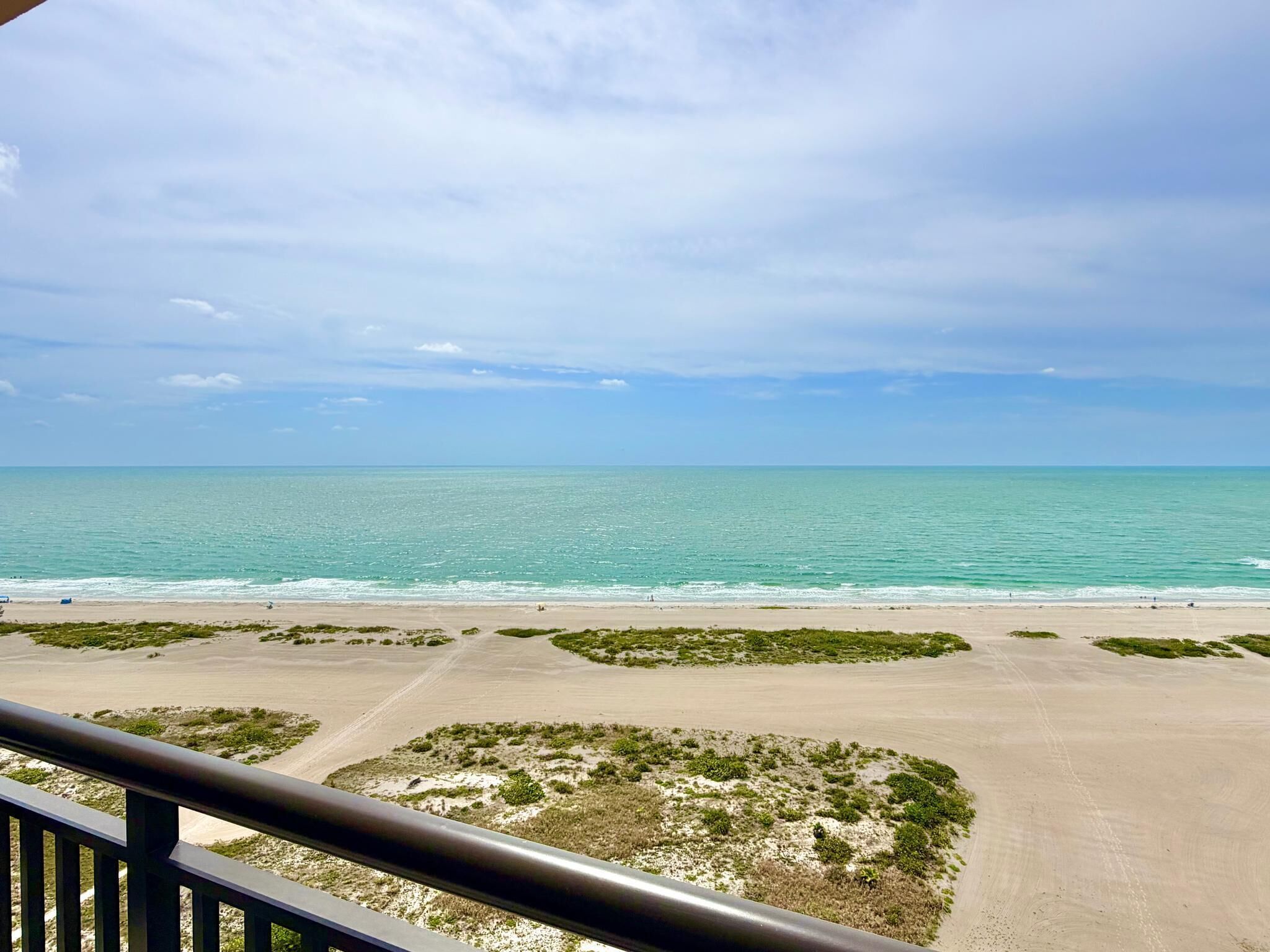 A beach view from a balcony with a railing, clear sky, and calm sea.