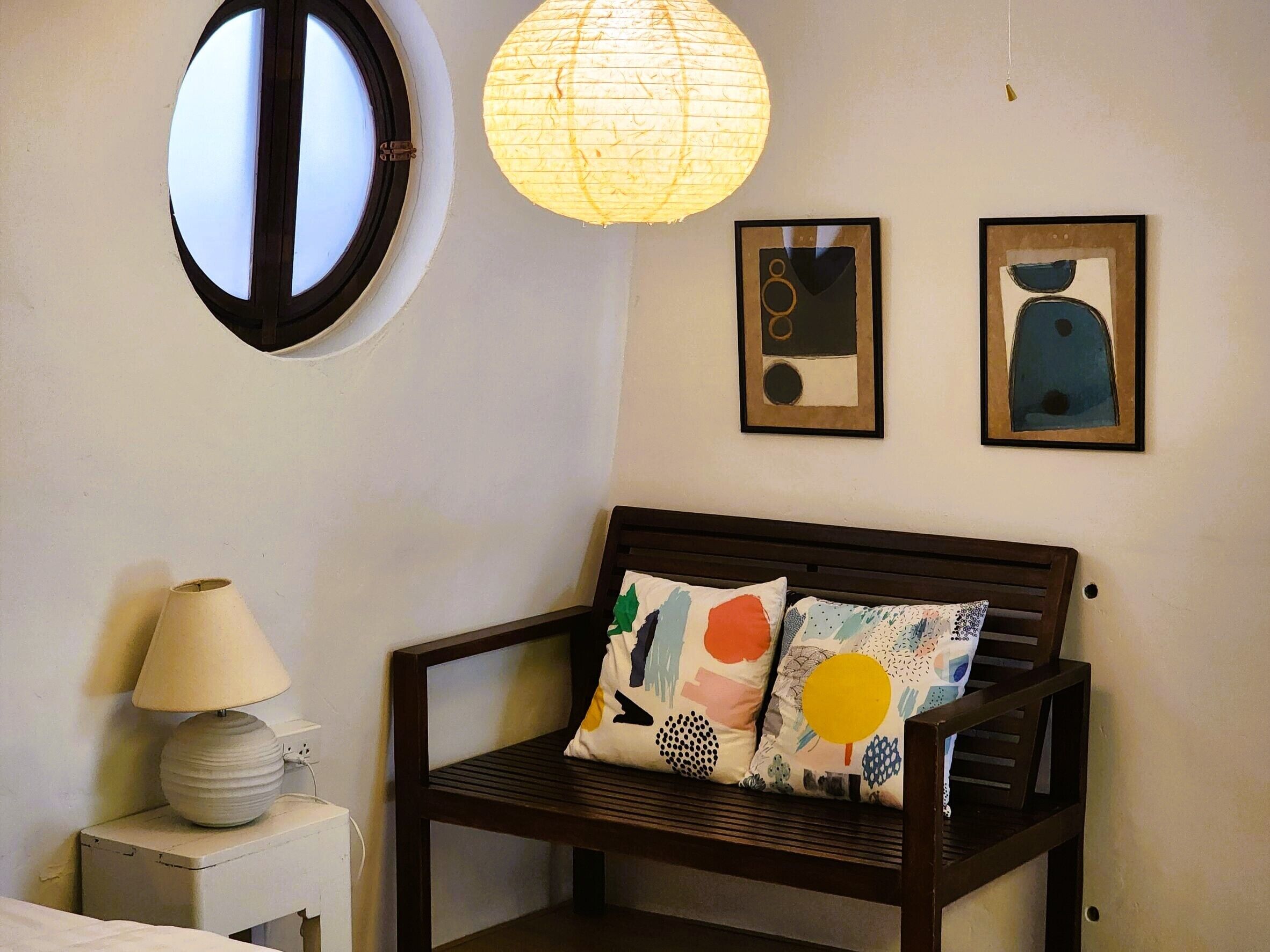 Una habitación con un banco de madera, dos almohadas con diseños abstractos, una lámpara y dos cuadros en la pared.
