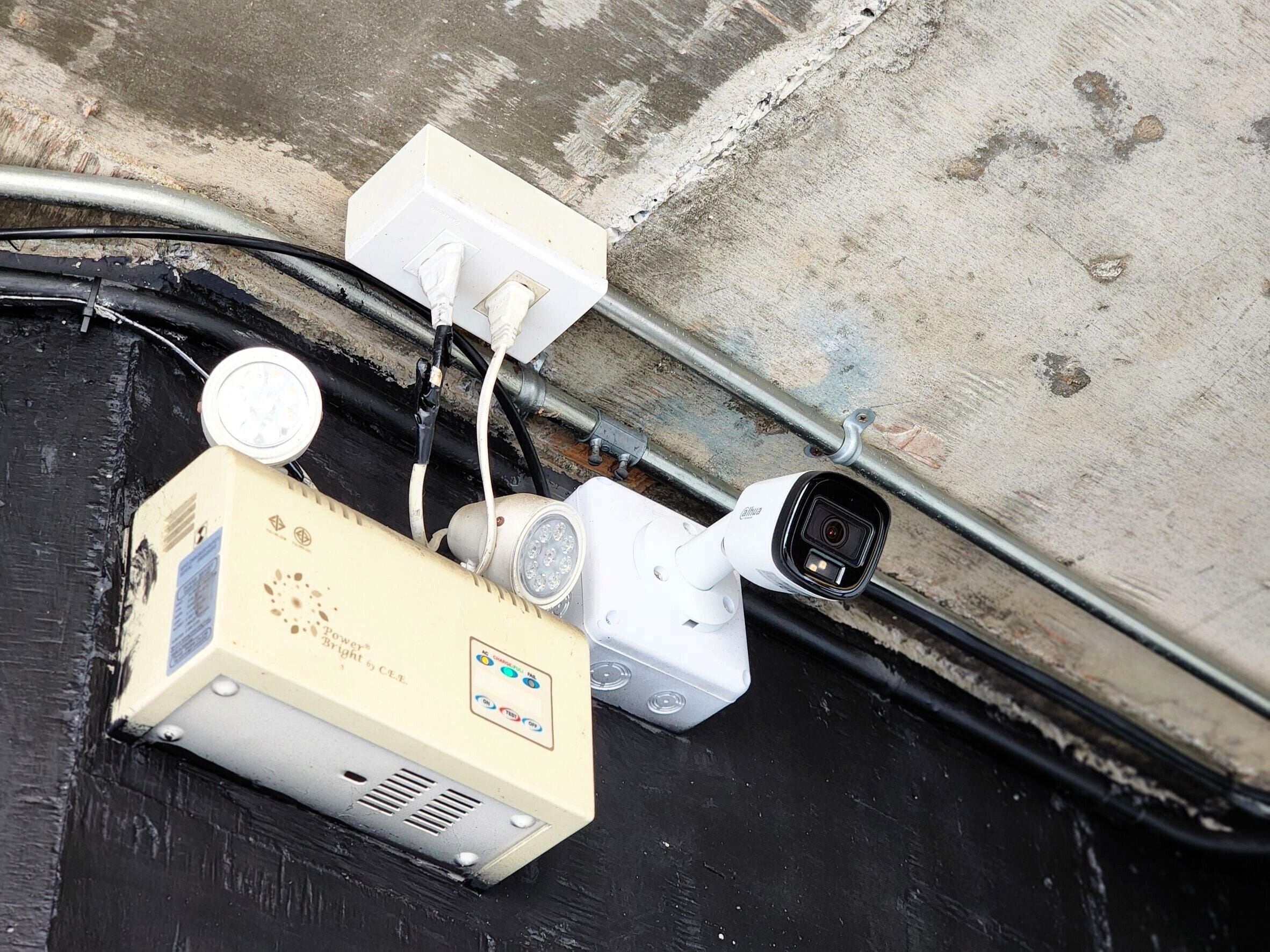 Una cámara de seguridad, un enchufe y un dispositivo con la etiqueta 'Power Bright' sobre una superficie de concreto.