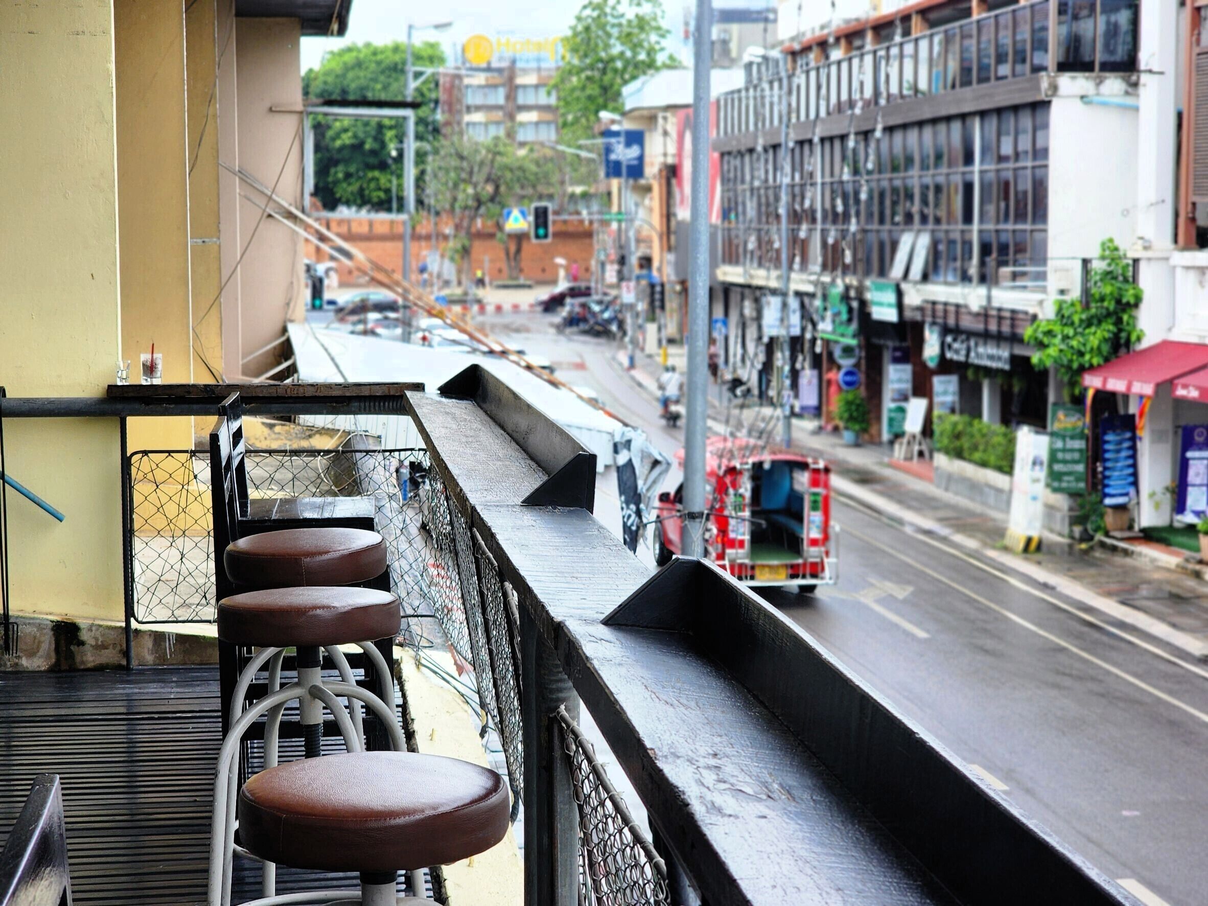 Un balcon doté d’une rambarde en métal, donnant sur une rue animée où circulent des véhicules et s’élèvent des immeubles.
