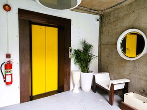 A room with a yellow door, a white wall-mounted mirror, a potted plant, a white chair, and a fire extinguisher. - MOD HOTEL CHIANG MAI (Chiang Mai)