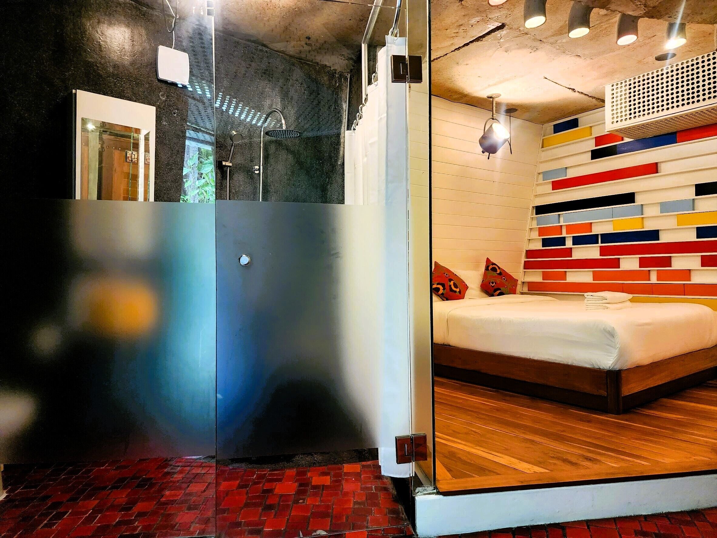 A modern hotel room with a glass partition, a shower area, a bed with colorful wall panels, and a wooden floor.