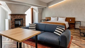 A modern hotel room with a brick fireplace, a large bed with a wooden headboard, a grey sofa with striped pillows, and a wooden table.