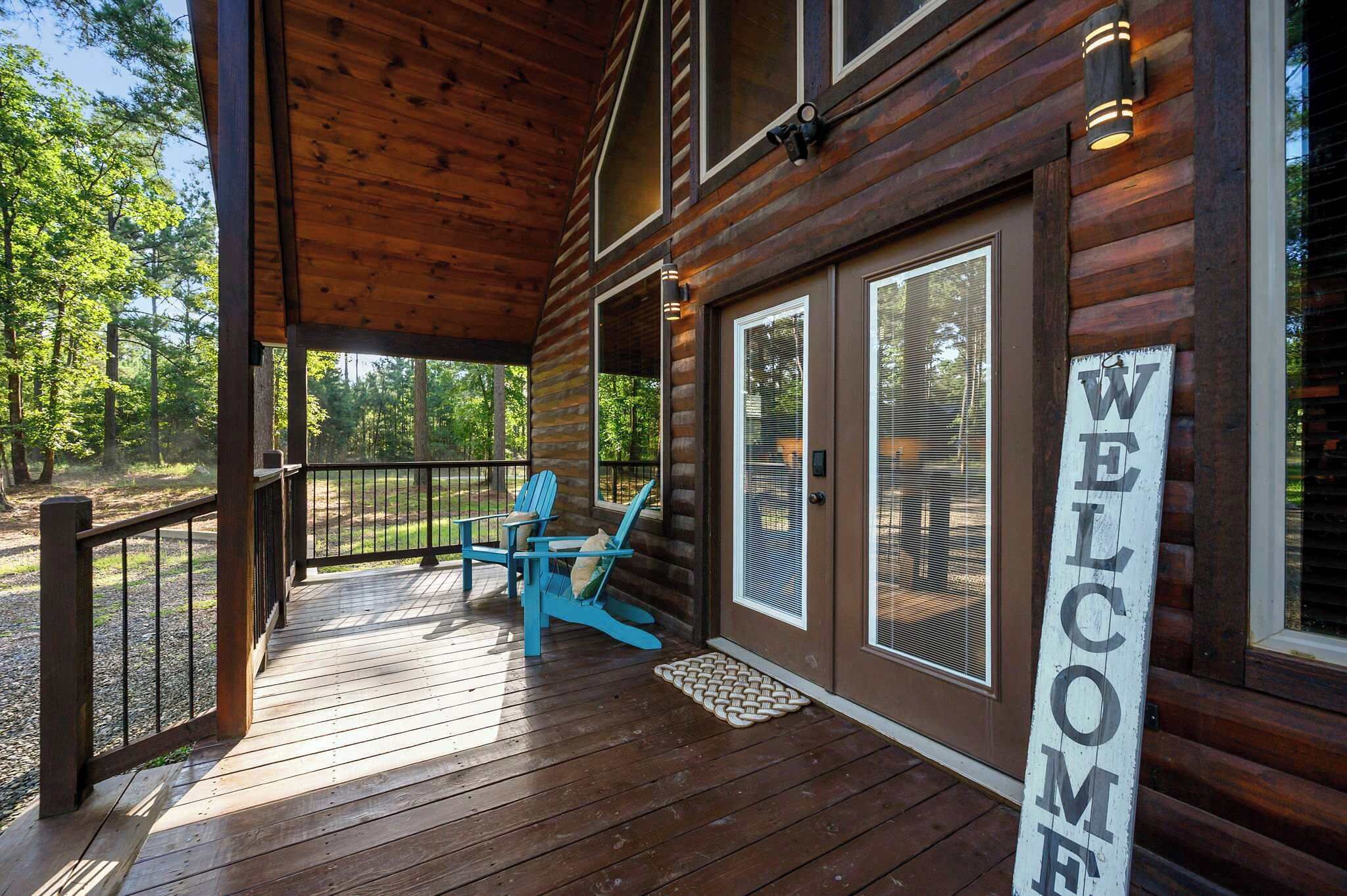 A wooden cabin with a porch, a welcome sign, and two blue Adirondack chairs.