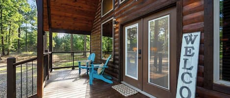 A wooden cabin with a porch, a welcome sign, and two blue Adirondack chairs.