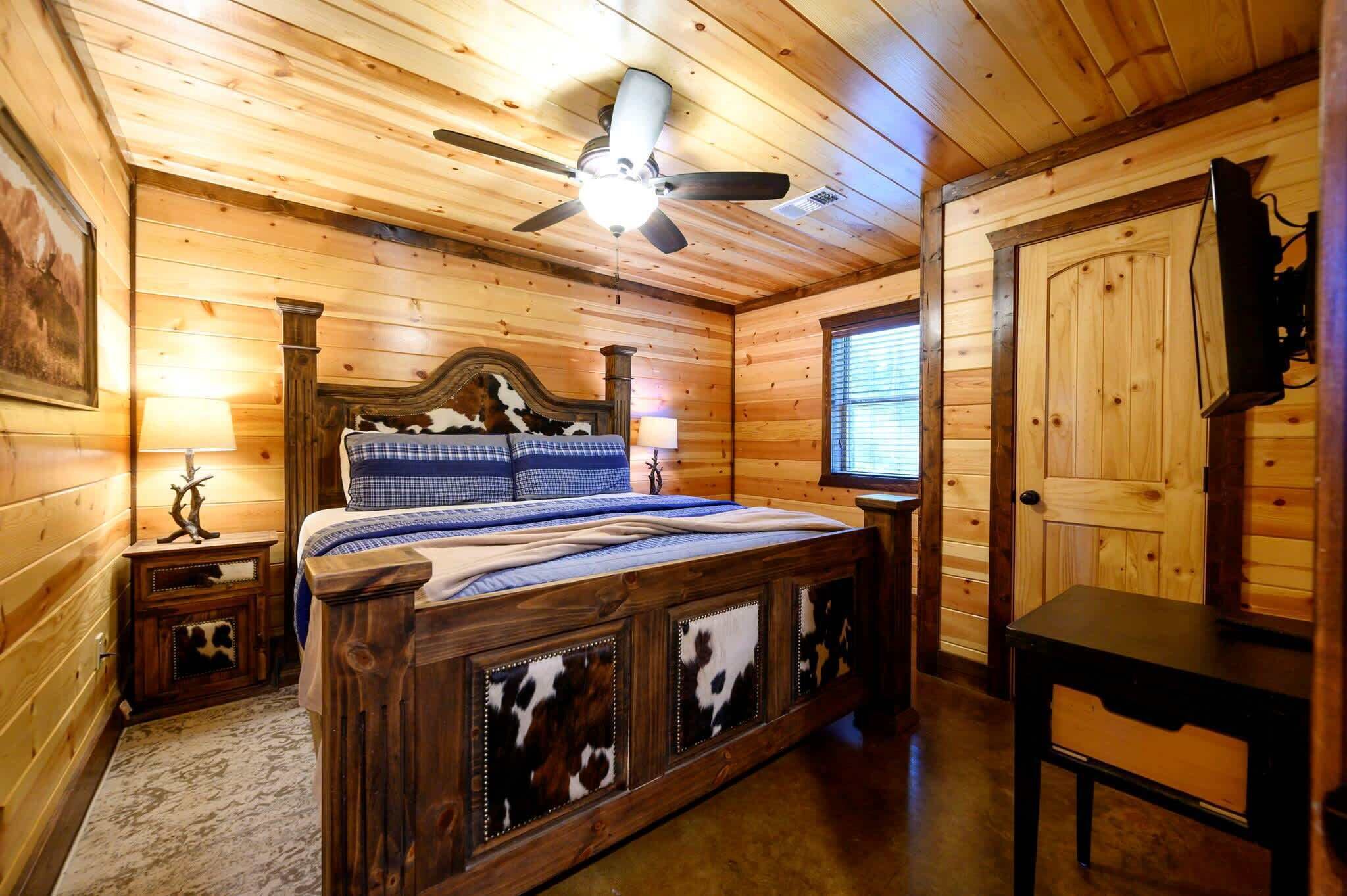 A wooden cabin bedroom with a bed, a ceiling fan, a nightstand, a lamp, a door, and a small table.