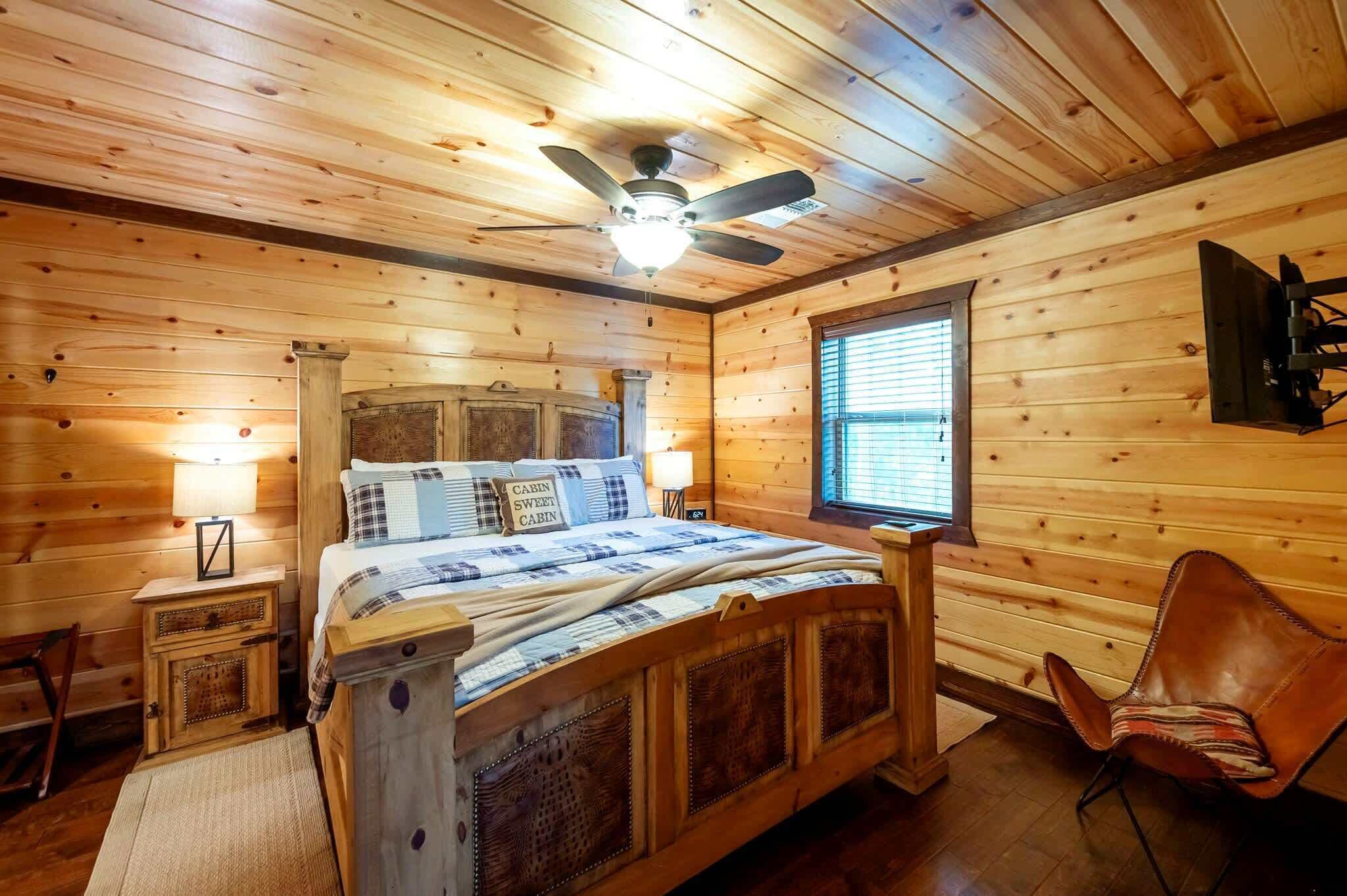A wooden cabin bedroom with a bed, a chair, a nightstand, a lamp, and a ceiling fan.