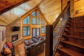 A cozy cabin interior with a stone fireplace, a wooden staircase, and large windows. - Over Yonder Inn (Broken Bow)