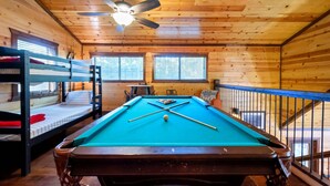 Een pooltafel in een houten kamer met stapelbedden en een plafondventilator.