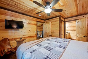 Een houten kamer met een bed, een plafondventilator, een tv aan de muur en een deur naar een andere kamer.
