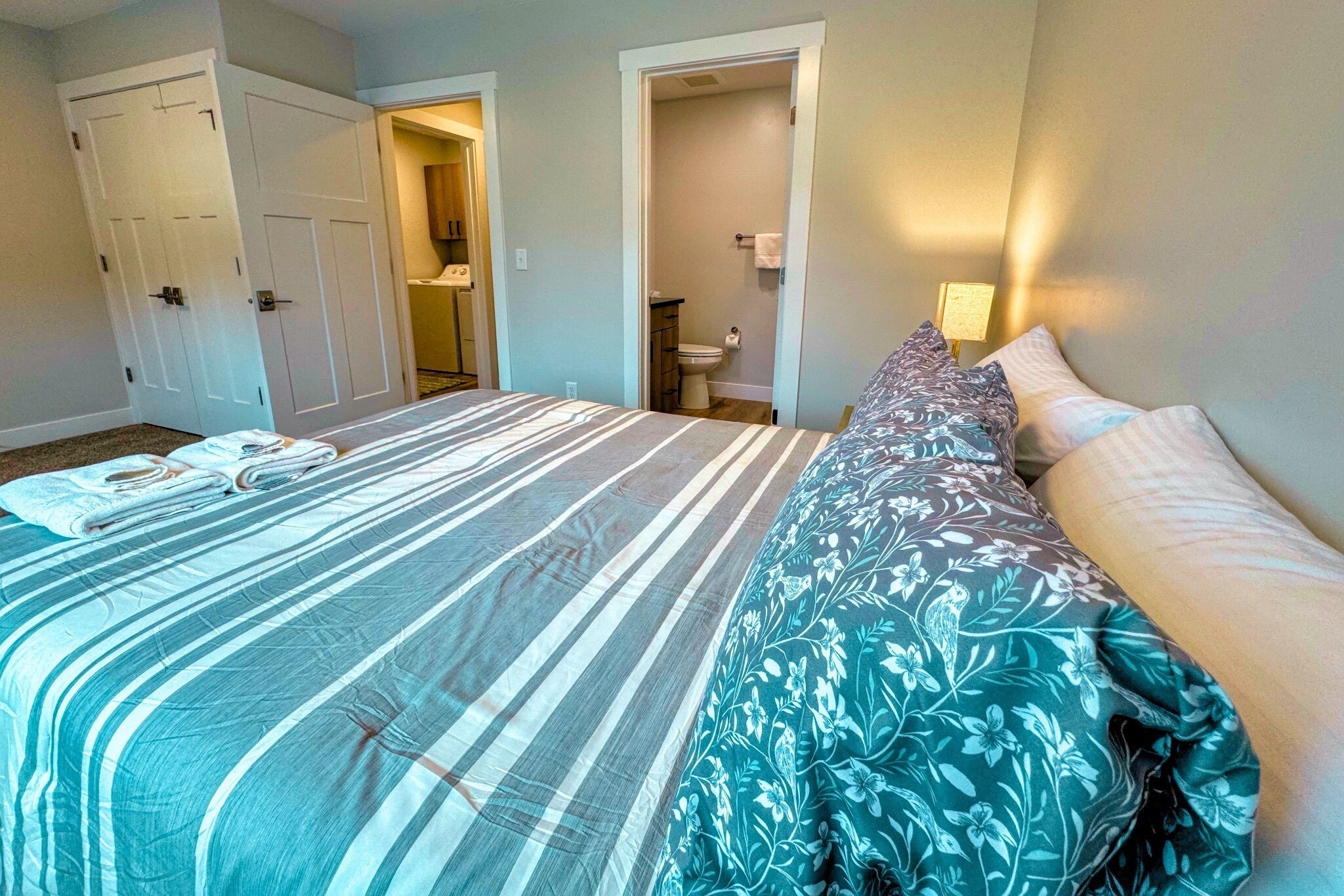 A bedroom with a bed, a nightstand, a door, and a bathroom visible through an open door.