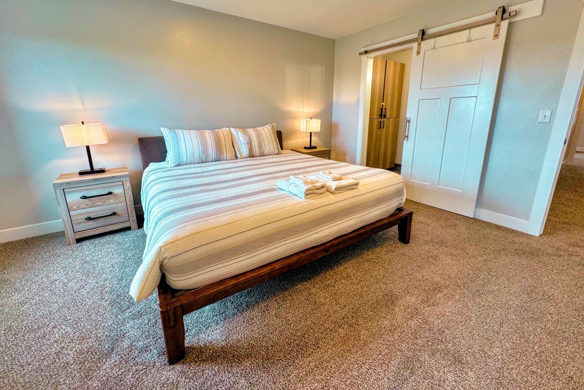 A neatly made bed with a striped bedspread and pillows, a bedside table with a lamp, and a wardrobe with sliding doors.