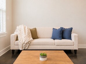 Un sofá beige con una manta blanca de punto y dos almohadas, una verde y otra azul, frente a una pared lisa. Delante del sofá, una mesa de centro de madera con una pequeña planta en maceta.