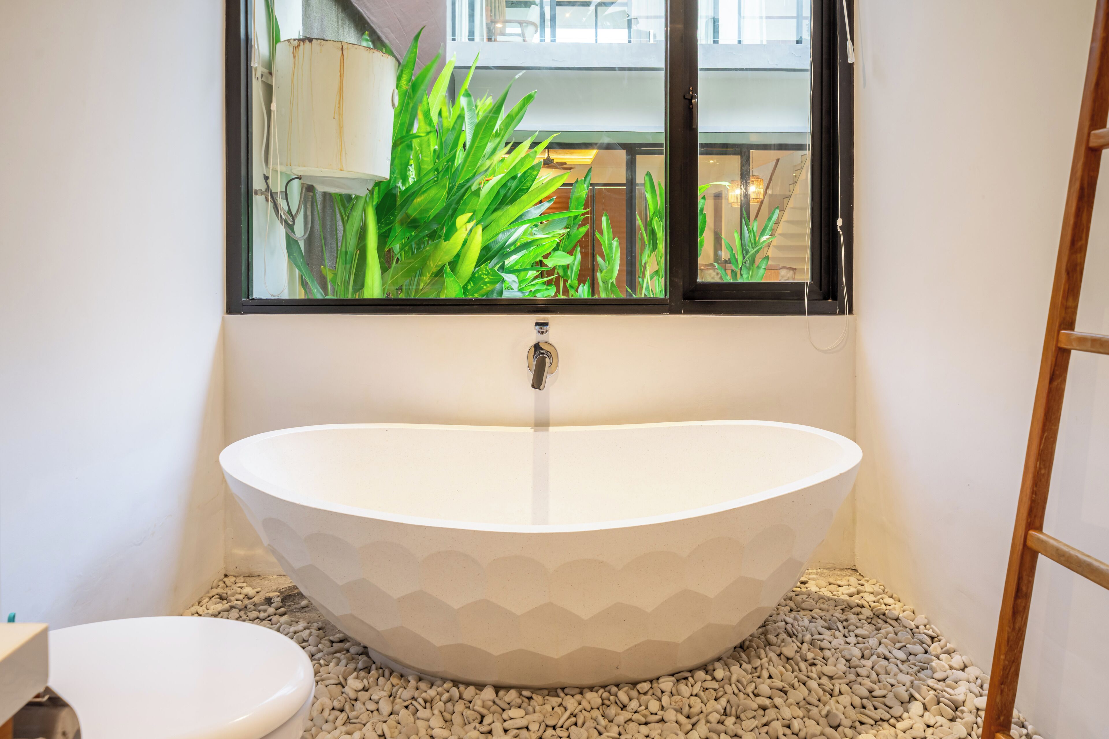 A modern bathroom with a freestanding bathtub, a toilet, and a window with a view of greenery.