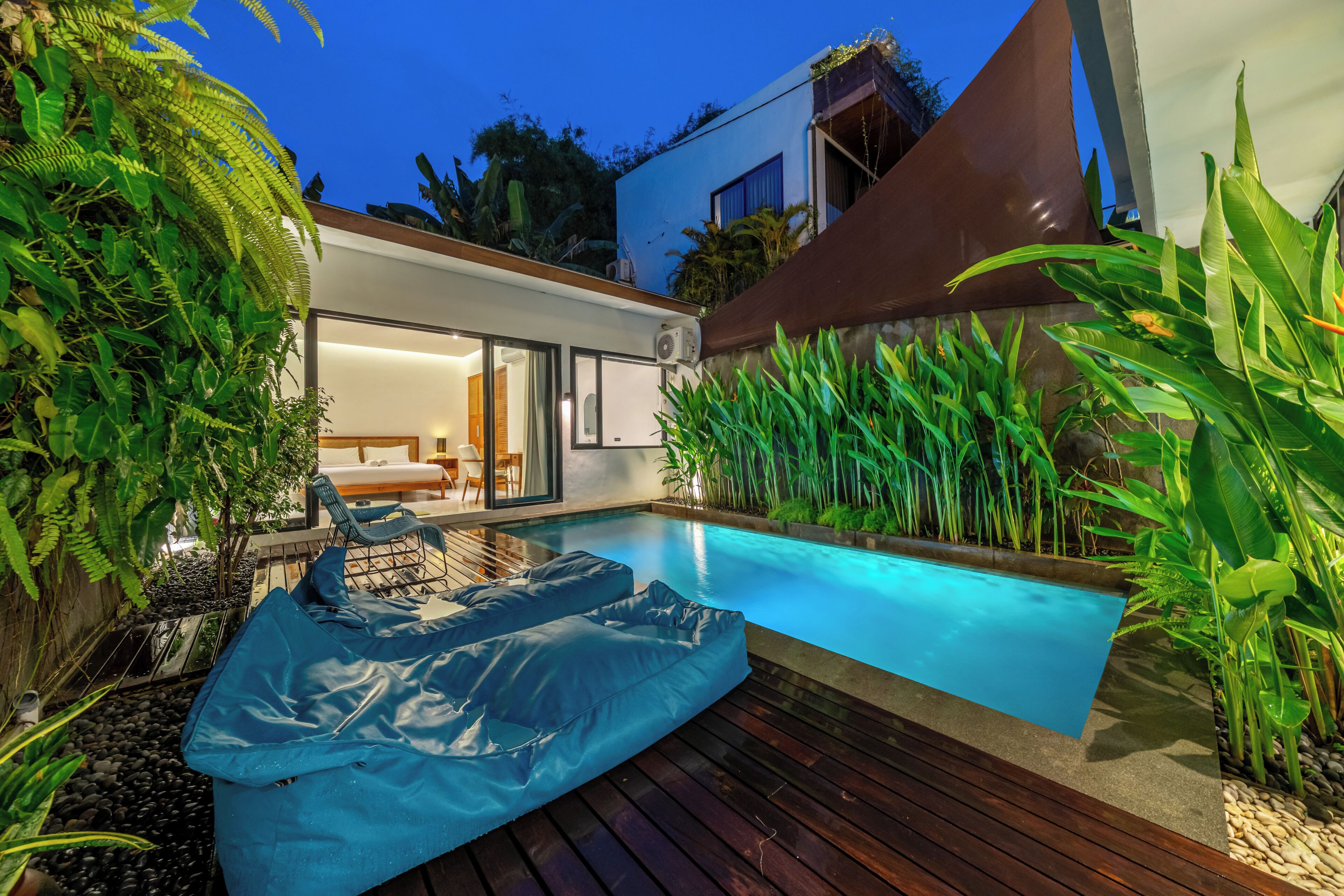 A modern outdoor pool area with a wooden deck, a blue inflatable lounge, and lush tropical plants.