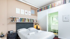 Un dormitori ben ordenat amb un llit, taules de nit, una prestatgeria i un bany visible a través d'una porta oberta.