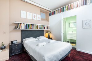 Ein ordentlich eingerichtetes Schlafzimmer mit einem Bett, Nachttischen, einem Bücherregal und einem durch eine offene Tür einsehbaren Badezimmer.