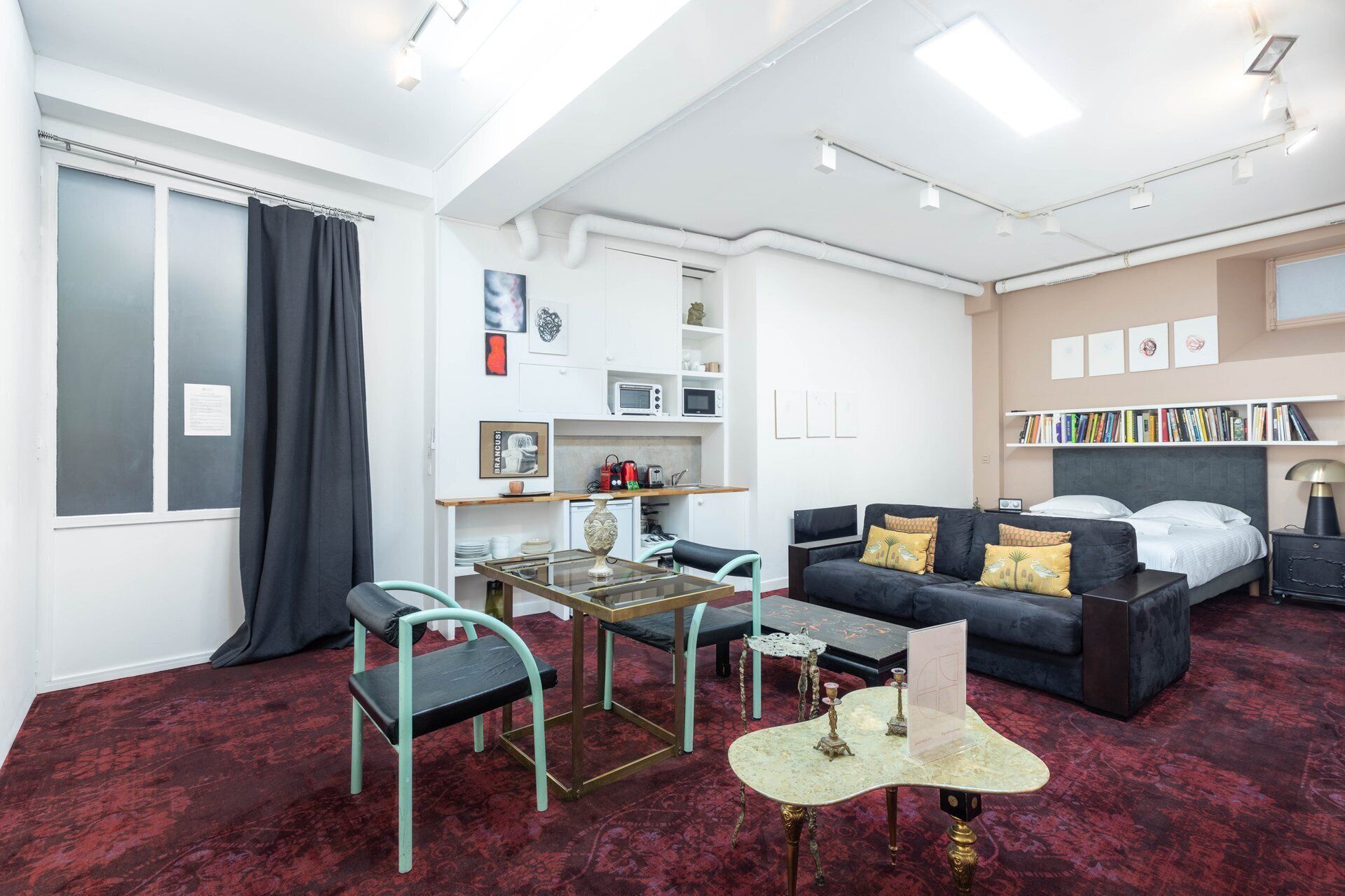 A modern hotel room with a bed, a sofa, a dining table, and a kitchenette.