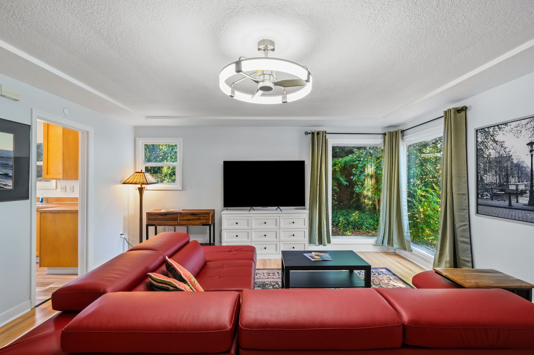 A modern living room with a large sofa, a flat-screen TV, a ceiling fan, and a view of greenery through large windows.