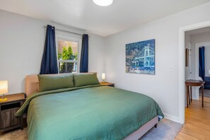 A bedroom with a large bed, a window with curtains, a nightstand with a lamp, and a painting on the wall. - Freshly renovated home with brand-new appliances, a new big-screen TV, refinished hardwood floors, and all-new furniture throughout. Sink into the plush leather sectional (pull-out queen sleeper) or dine at the six-seat table in the garden. (Vancouver)