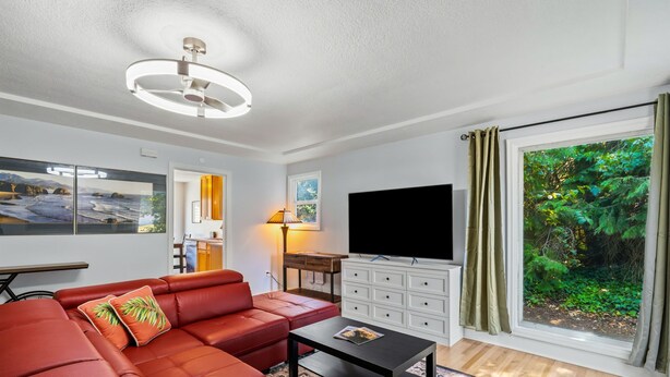 A modern living room with a sectional sofa, a flat-screen TV, a coffee table, and a large window with a view of greenery. - Freshly renovated home with brand-new appliances, a new big-screen TV, refinished hardwood floors, and all-new furniture throughout. Sink into the plush leather sectional (pull-out queen sleeper) or dine at the six-seat table in the garden. (Vancouver)