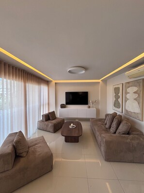 A modern living room with a flat-screen TV, two sofas, a coffee table, and a large window with curtains. - Crystal Green Signature Properties  (Athina)