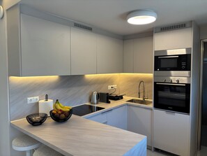 A modern kitchen with a built-in microwave, oven, and sink. There is a fruit bowl on the counter and a kettle. - Crystal Green Signature Properties  (Athina)