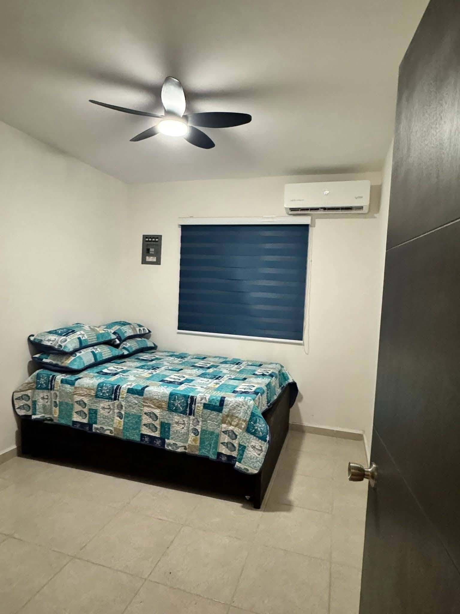 Een slaapkamer met een bed, een plafondventilator, een airconditioner, een raam met jaloezieën en een deur.