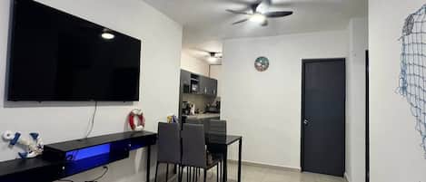 A modern living room with a flat-screen TV, a ceiling fan, a dining area with chairs, and a kitchen in the background.