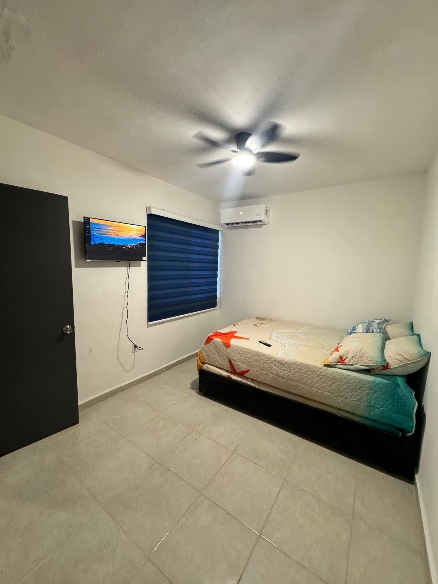 Une chambre à coucher comprenant un lit, un ventilateur de plafond, un climatiseur, une télévision et une fenêtre avec des stores.