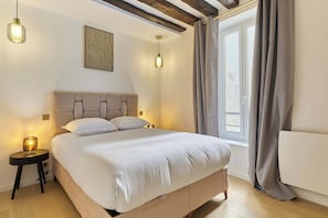 Ein Schlafzimmer mit einem Bett, Nachttischlampen, einem Fenster mit Vorhängen und einem Holzbalken an der Decke.