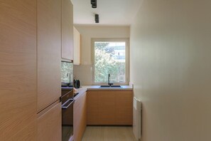 Eine kleine Küche mit Holzschränken, einem eingebauten Ofen, einem Fenster mit Blick auf Häuser und einer weißen Arbeitsplatte.