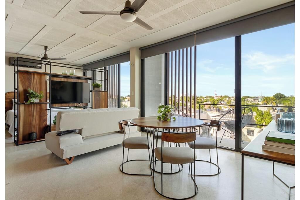 A modern living room with a sofa, a dining table with chairs, a TV, and large windows offering a city view.