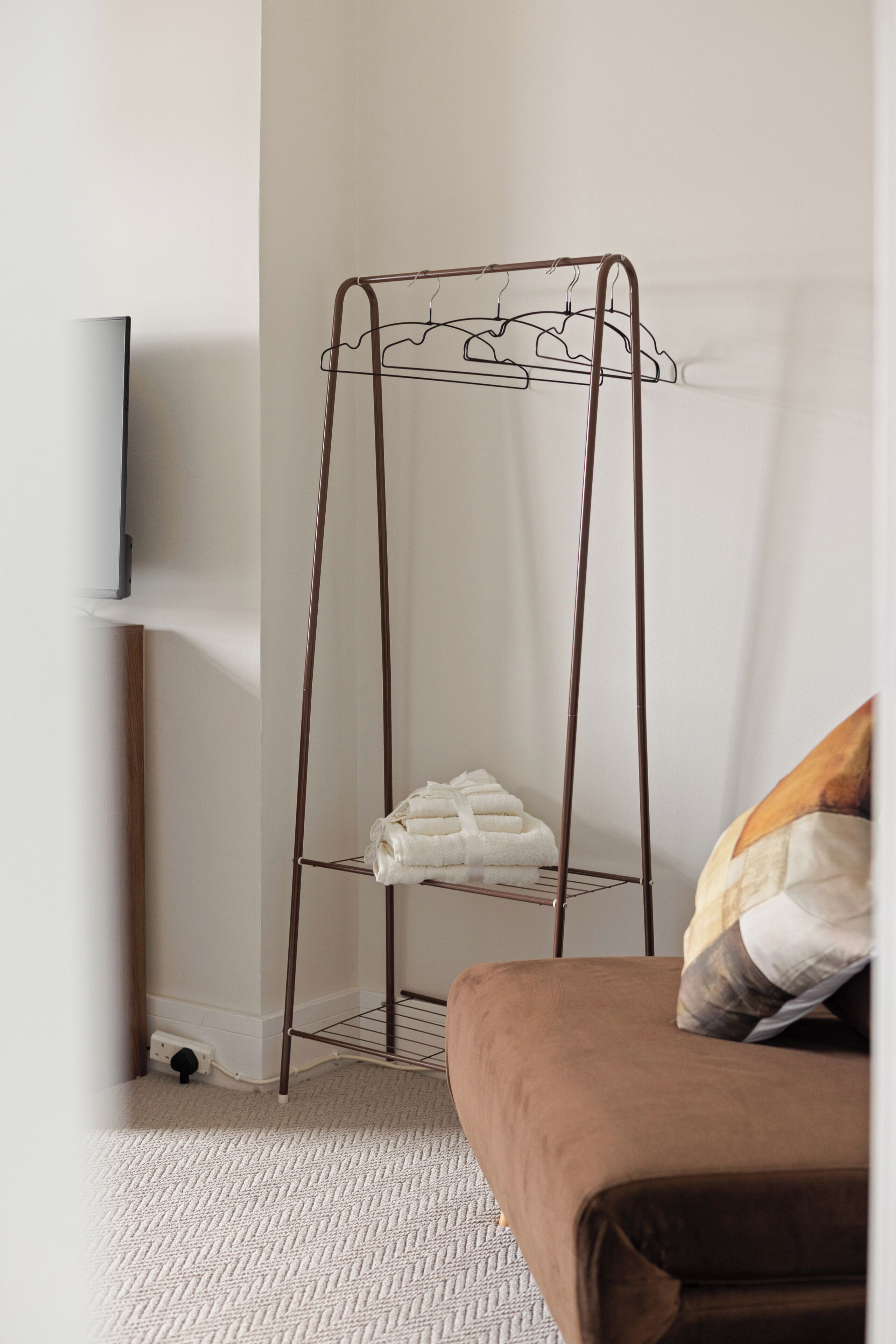 A room with a metal wire shelf unit against a wall, a television mounted on the wall, and a bed with a brown cover.