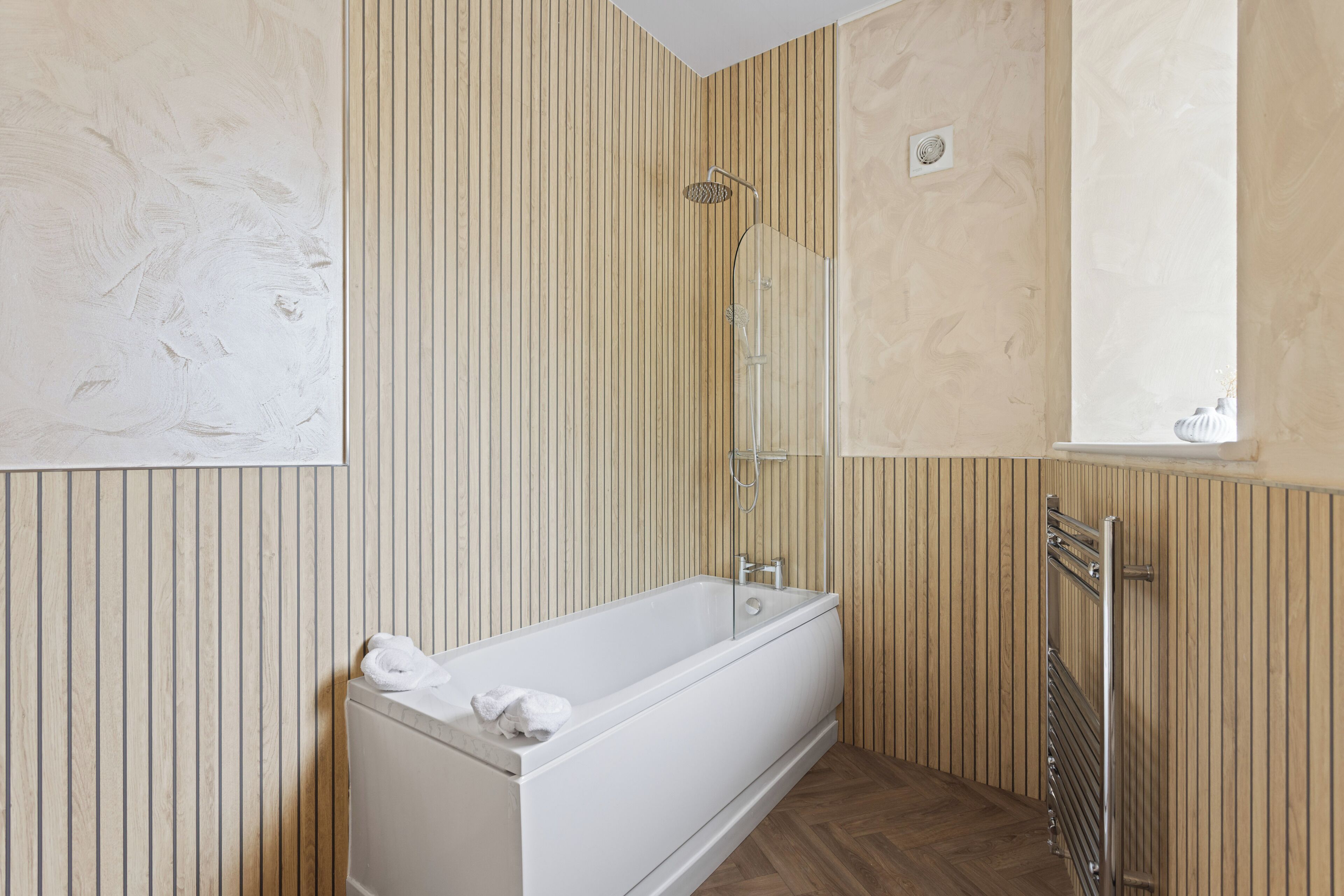 A bathroom with a white bathtub, wooden paneling, a mirror, and a window.