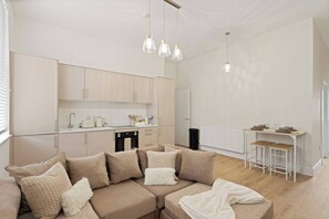A modern kitchen with white cabinets, a dining area with a table and bar stools, and a living room with a beige sectional sofa.
