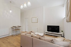 A modern living room with a beige sofa, a TV on a white stand, and a dining area with a white table and chairs. - Family-Friendly 3BR Home I Parking I Sleeps 5 I Broxburn (West Lothian)